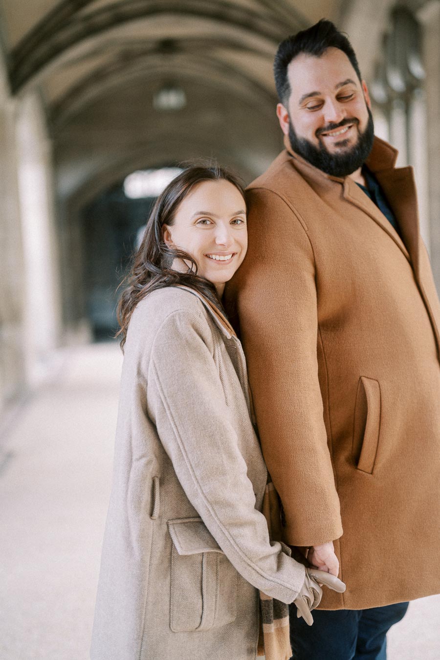A joyful couple in stylish winter coats holding hands in an elegant archway, exuding warmth and affection.