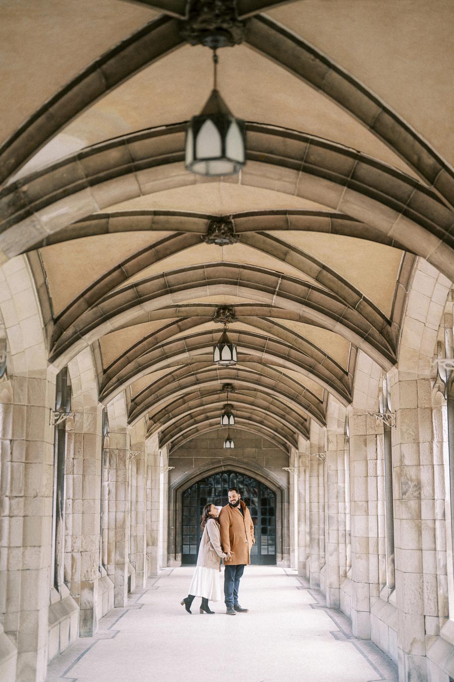 Couple walking in historic arched stone hallway, dressed in winter coats and smiling at each other, in an elegant architectural setting.