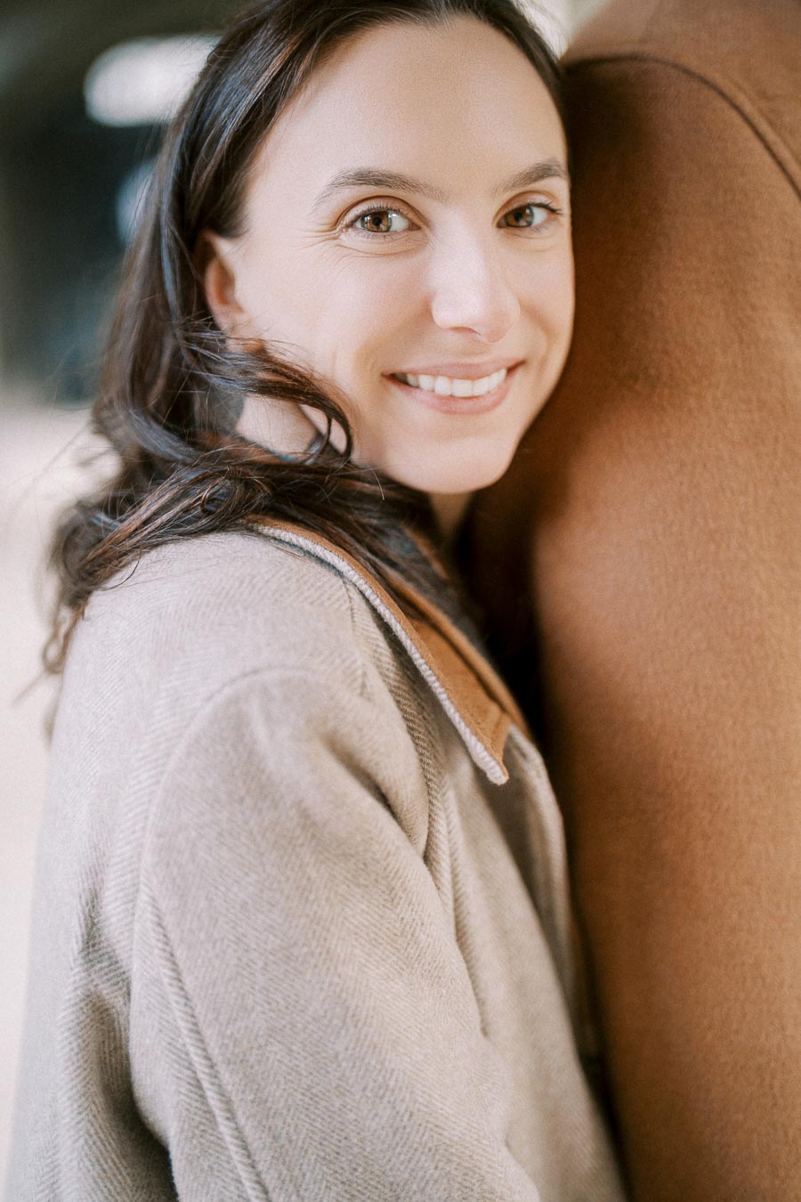 Smiling woman in a beige coat standing close to a person in a brown coat, looking directly at the camera with a blurred urban background.