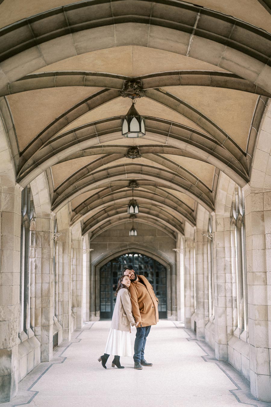 A couple standing under a historic stone archway, smiling and dressed warmly, within a beautifully lit corridor.