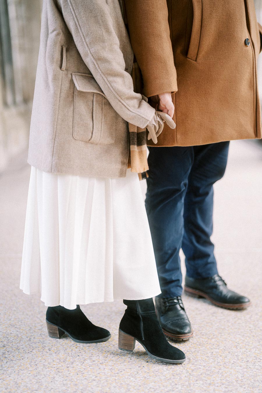 A couple holding hands wearing stylish winter coats, leather shoes, and boots, standing on a textured pavement, emphasizing fashion and seasonal attire.