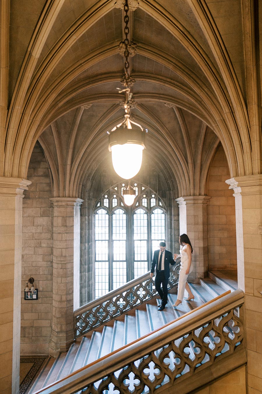 A couple elegantly walking down a grand, historic staircase with intricate stonework and a large arched window in a