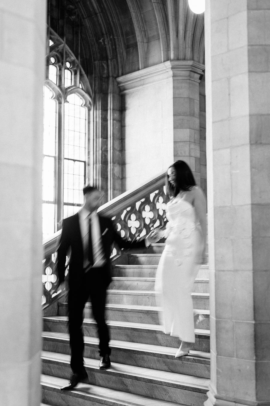 A couple in formal attire gracefully descends a grand staircase in a historic building, with gothic architecture and elegant