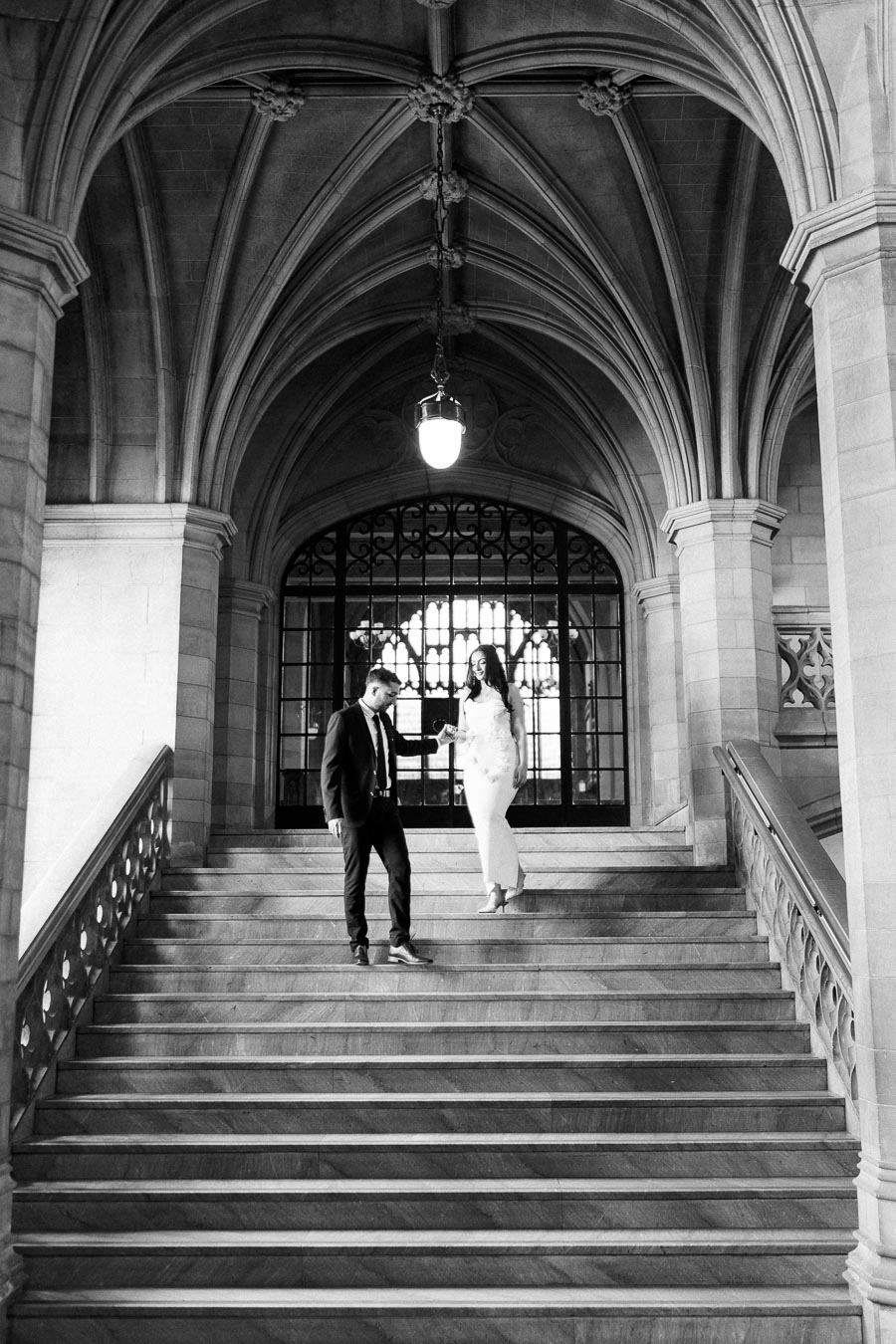 Black and white photo of a couple in formal attire walking down a grand stone staircase in a historic building with arched