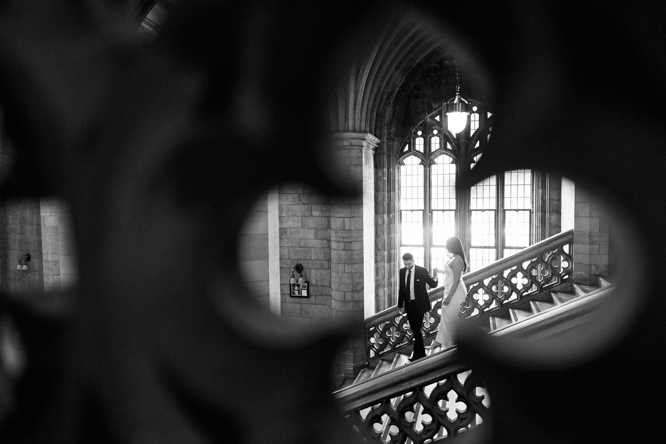 A couple descending an ornate staircase in a historic building, framed by intricate architectural details, with large leaded