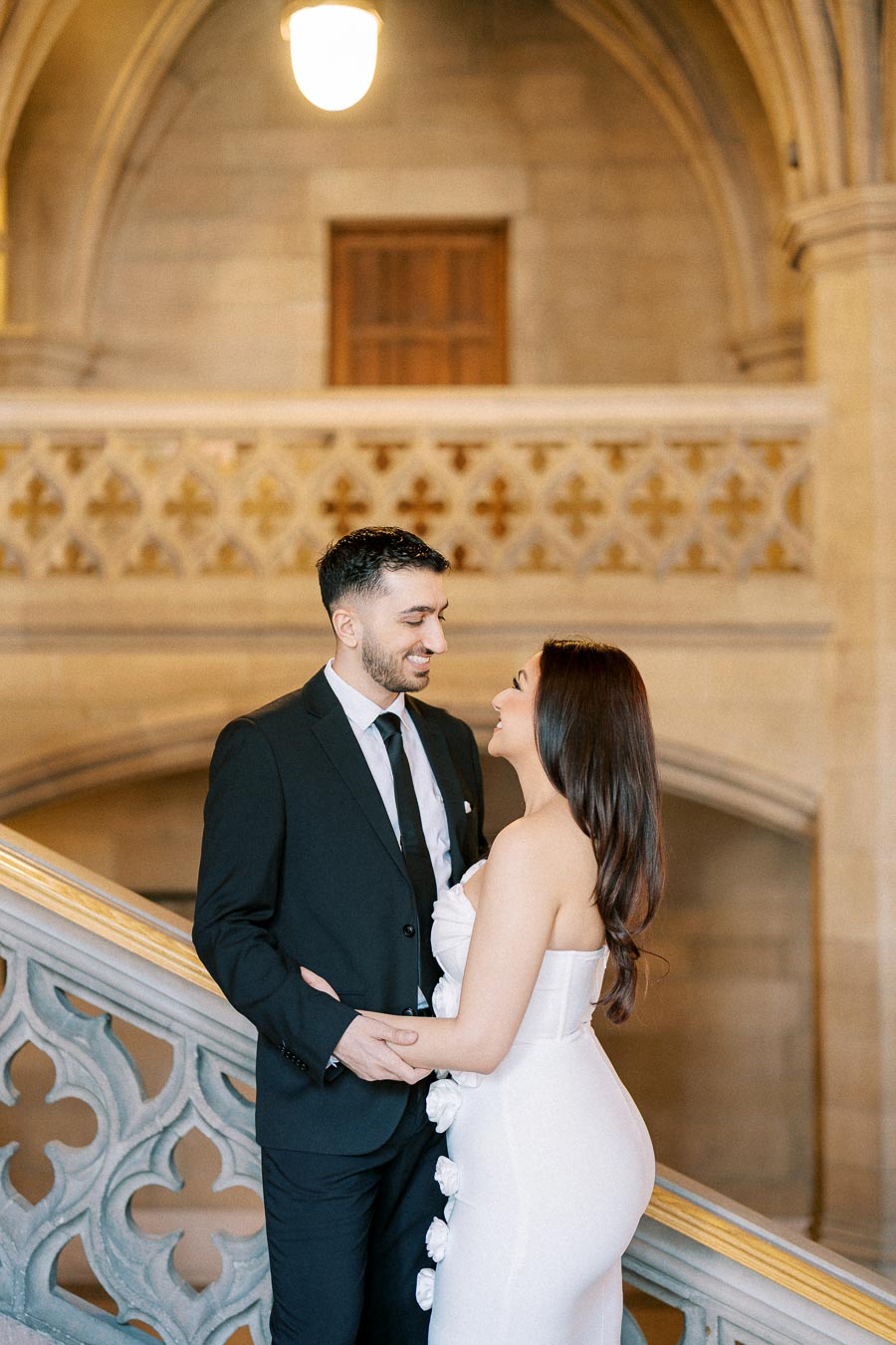 A couple dressed in formal attire shares a romantic moment on a grand staircase inside a historic building with ornate