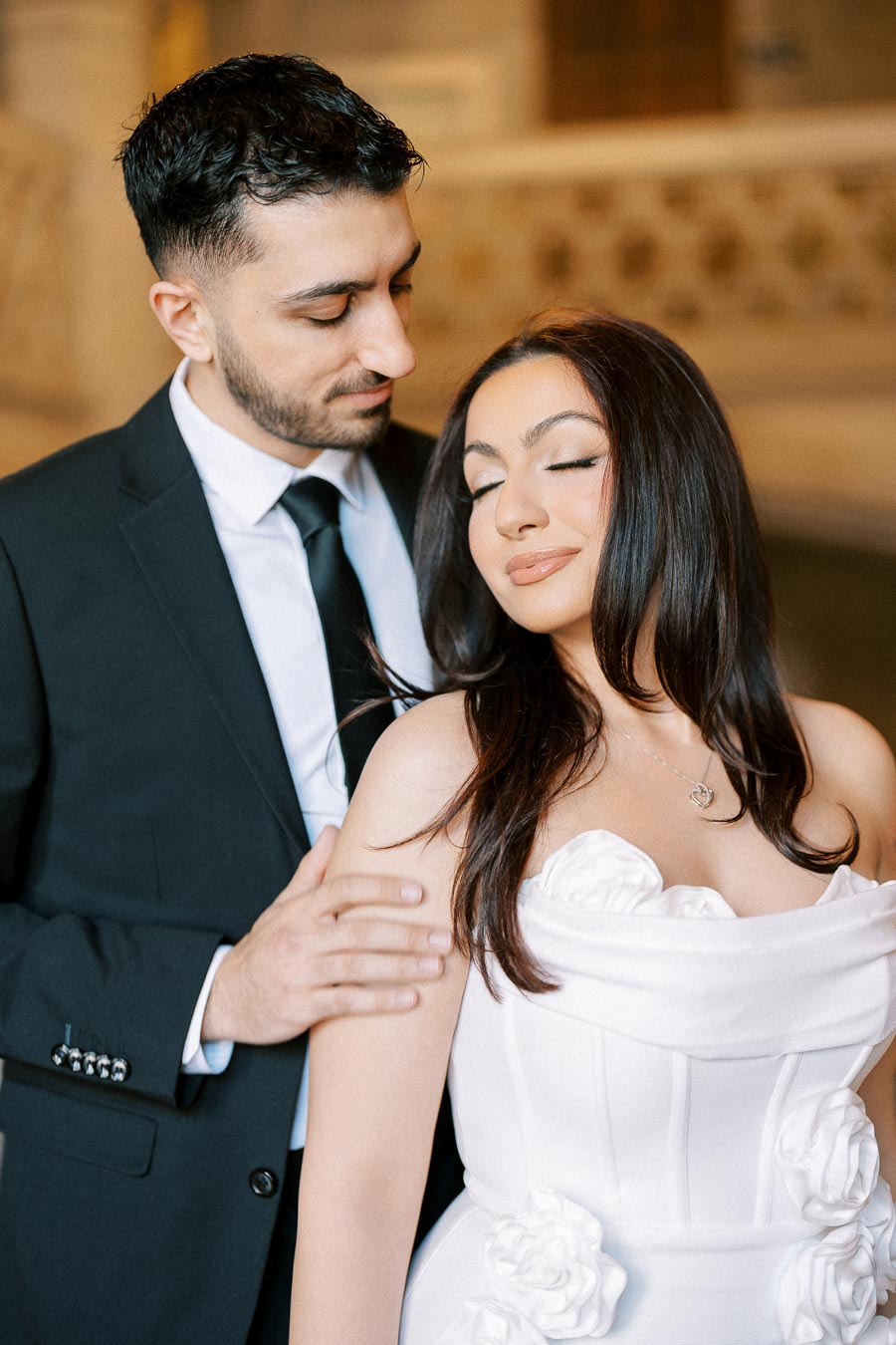 A couple embracing each other, with the woman wearing a white dress adorned with floral details and the man in a black suit,