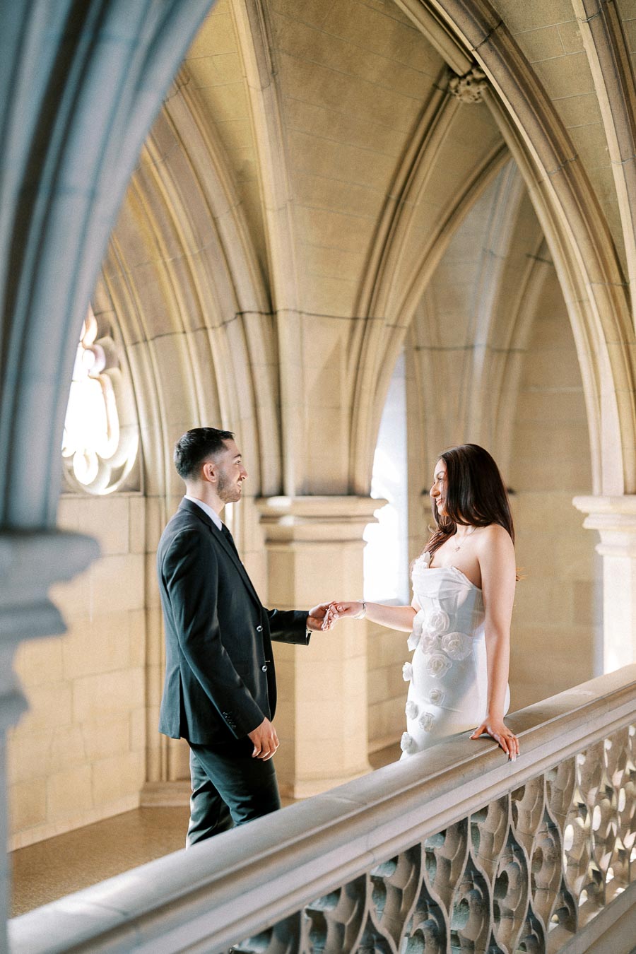 A couple in elegant attire holding hands in a beautifully lit, arched stone hallway.