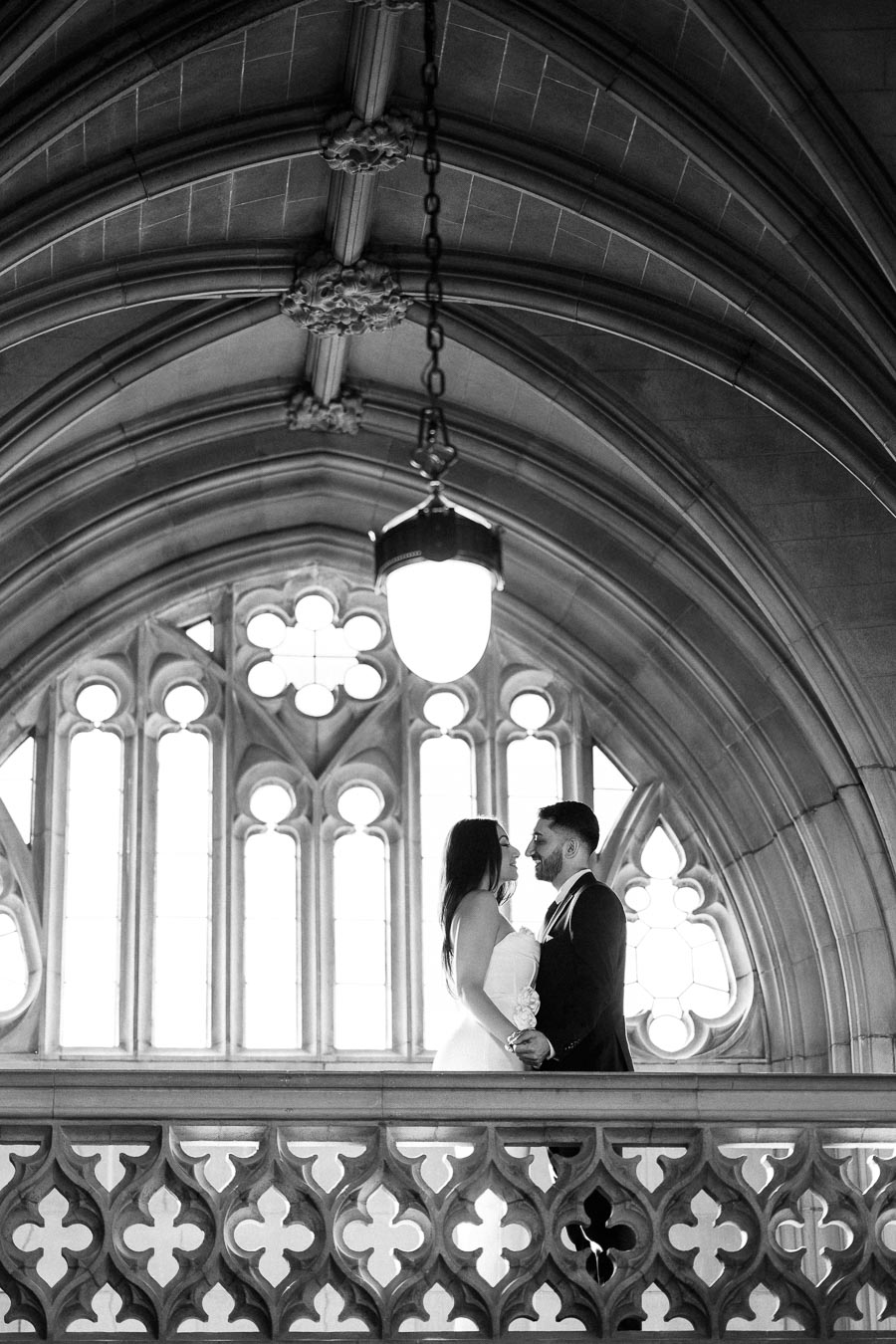 Black and white photo of a couple standing on a decorative balcony in a historic building, with arched ceilings and