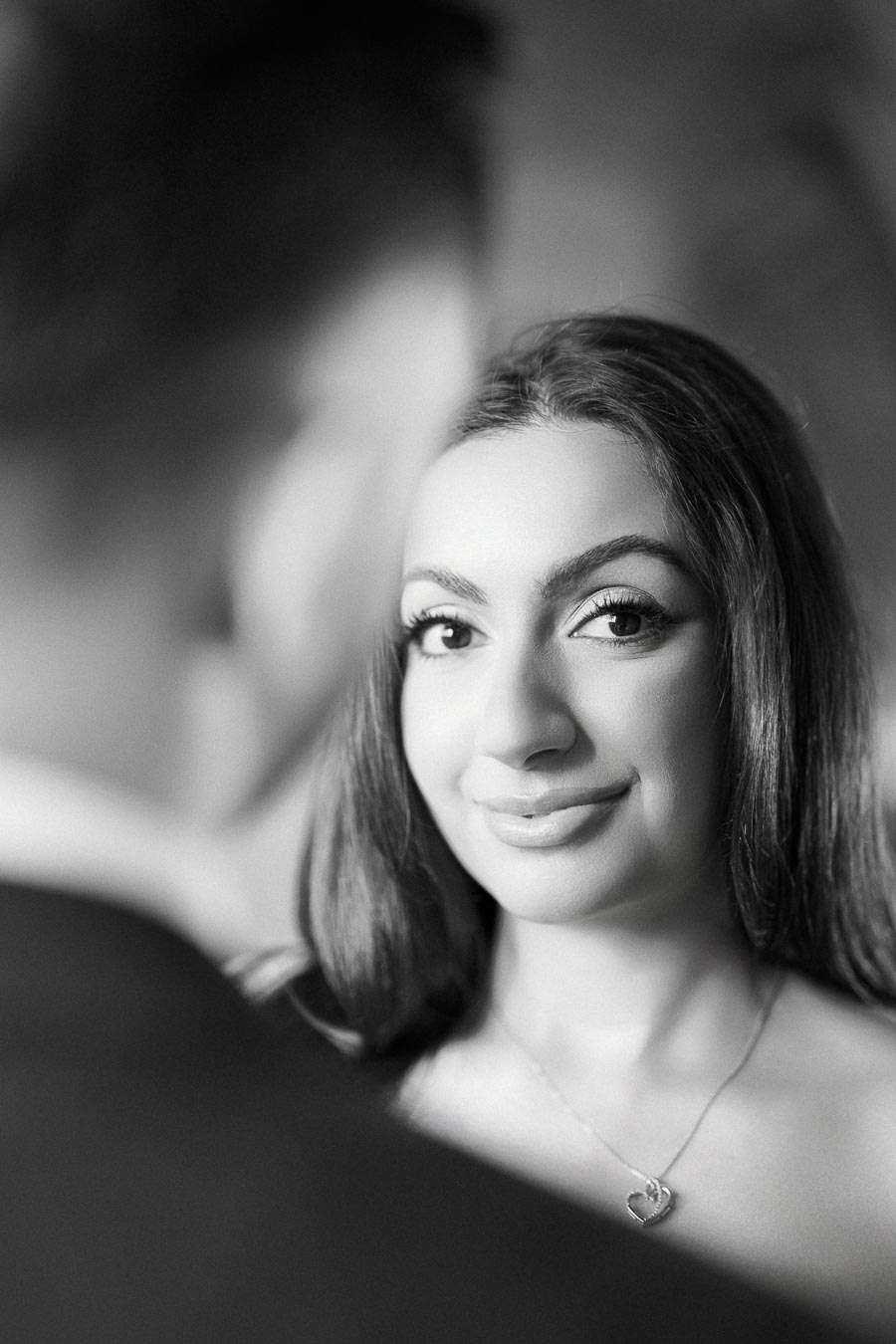 Black and white portrait of a smiling woman with long hair, wearing a heart-shaped necklace, looking towards the camera with
