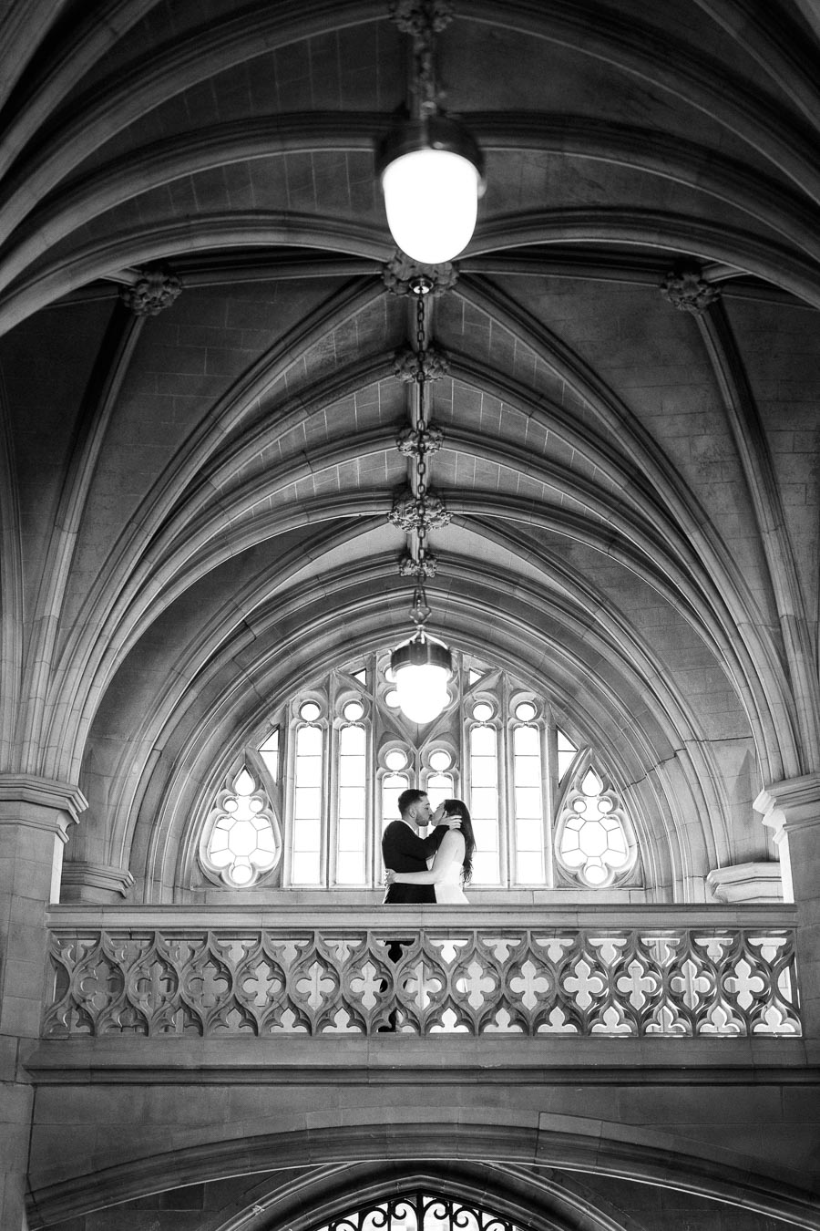 A couple embracing on a Gothic-style balcony under ornate arched ceilings, with intricate stonework and large windows in a