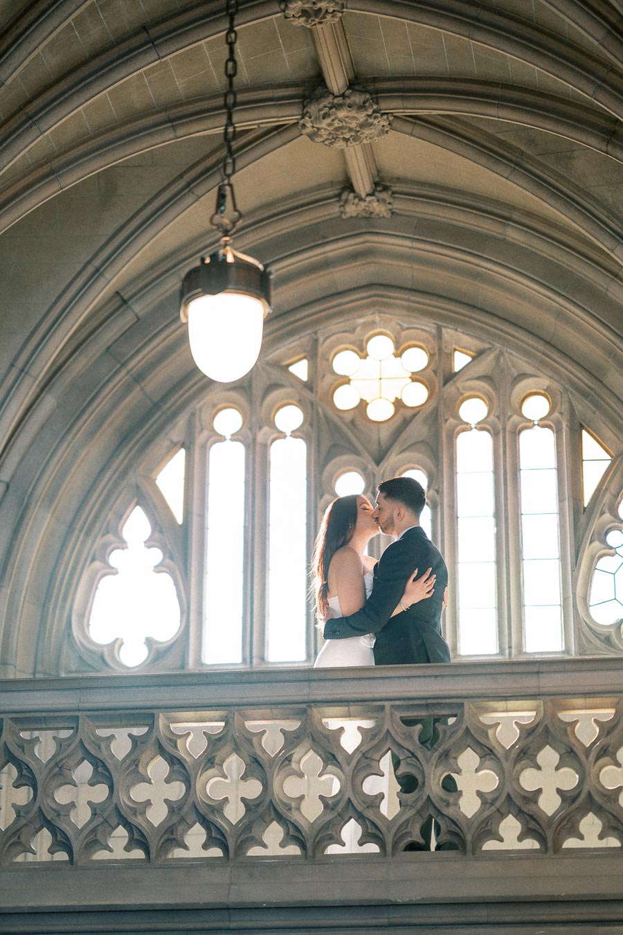 A couple embracing and kissing in an elegant, historic arched hallway with decorative stone patterns and large windows in
