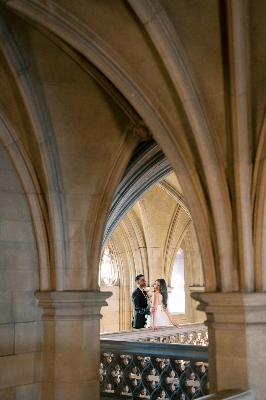 A couple standing on an ornate balcony, surrounded by Gothic archways and stone architecture, creates a romantic and elegant