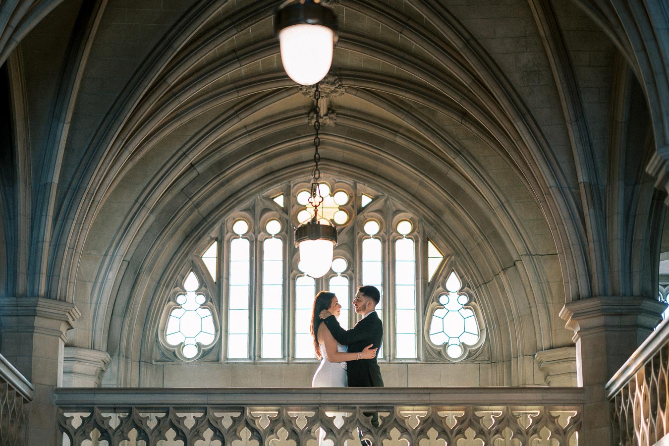 A couple embracing in an elegant gothic-style hall with arched windows and intricate architectural details.
