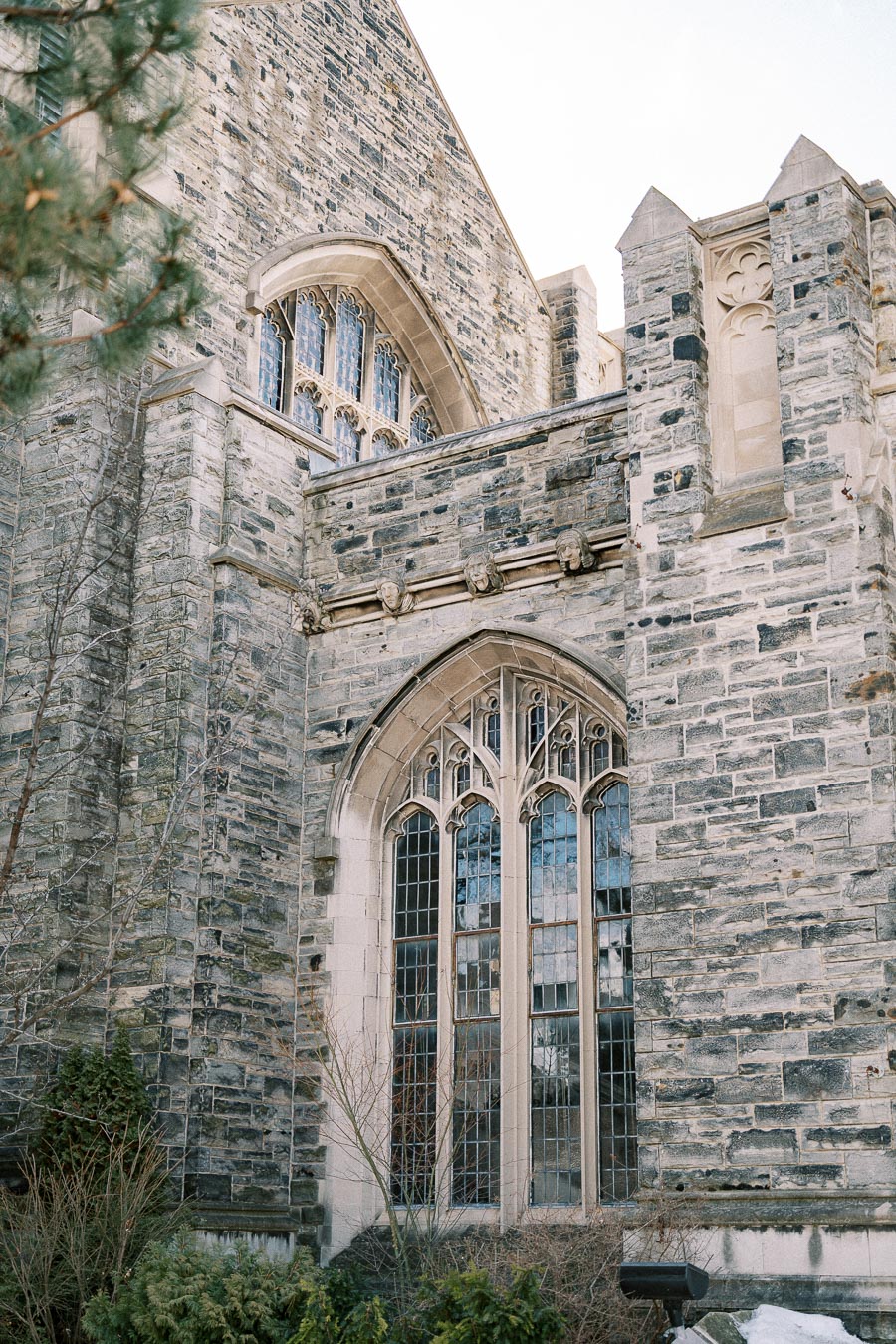 Gothic-style stone building facade with large arched windows and intricate architectural details, surrounded by greenery.