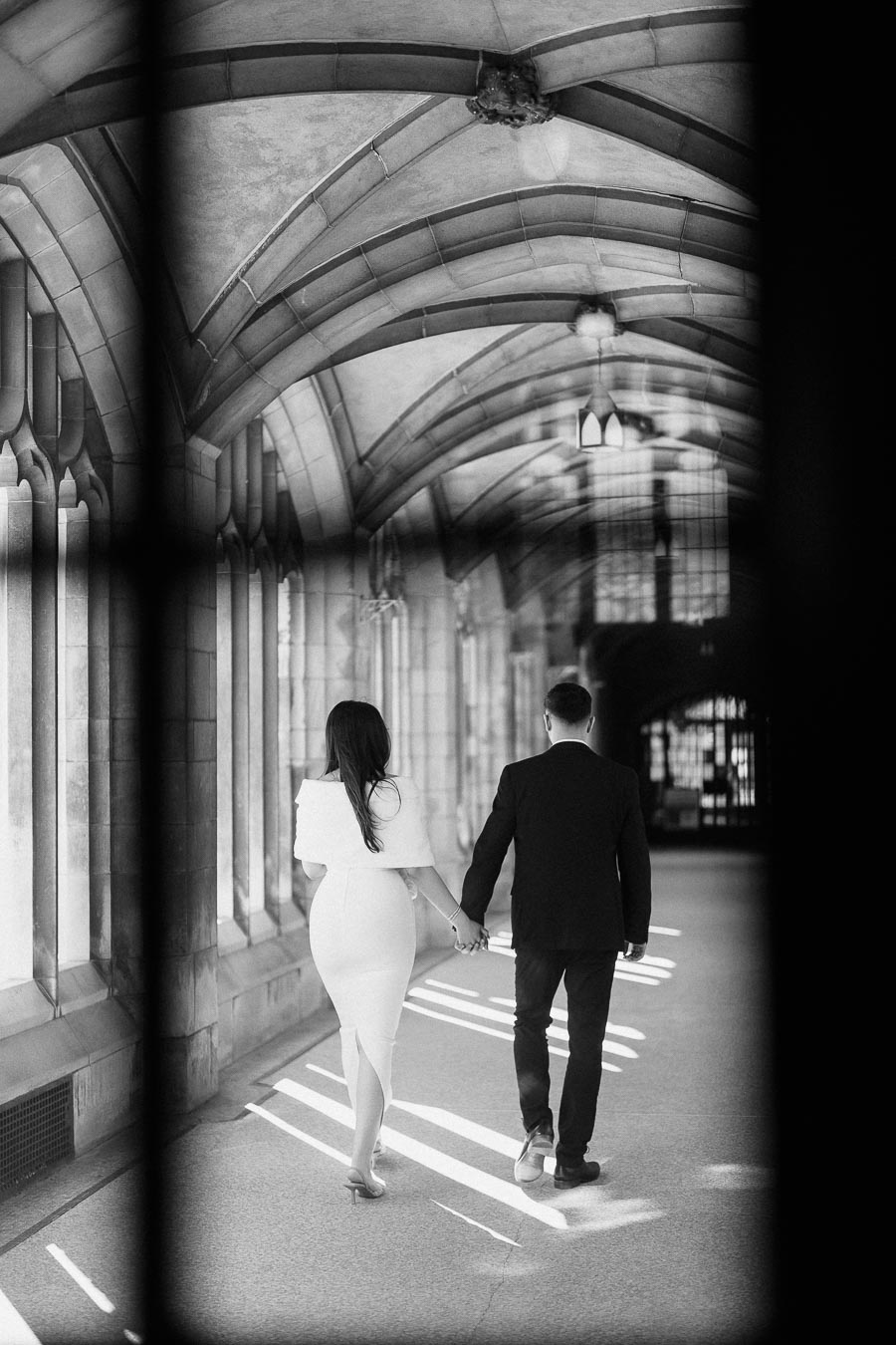 A couple holding hands walks through a beautifully arched corridor with stone architecture, casting shadows on the ground,