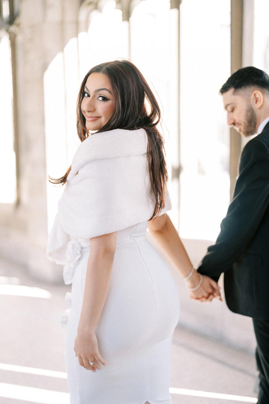 Elegant couple holding hands in a bright setting, woman in white dress smiling back
