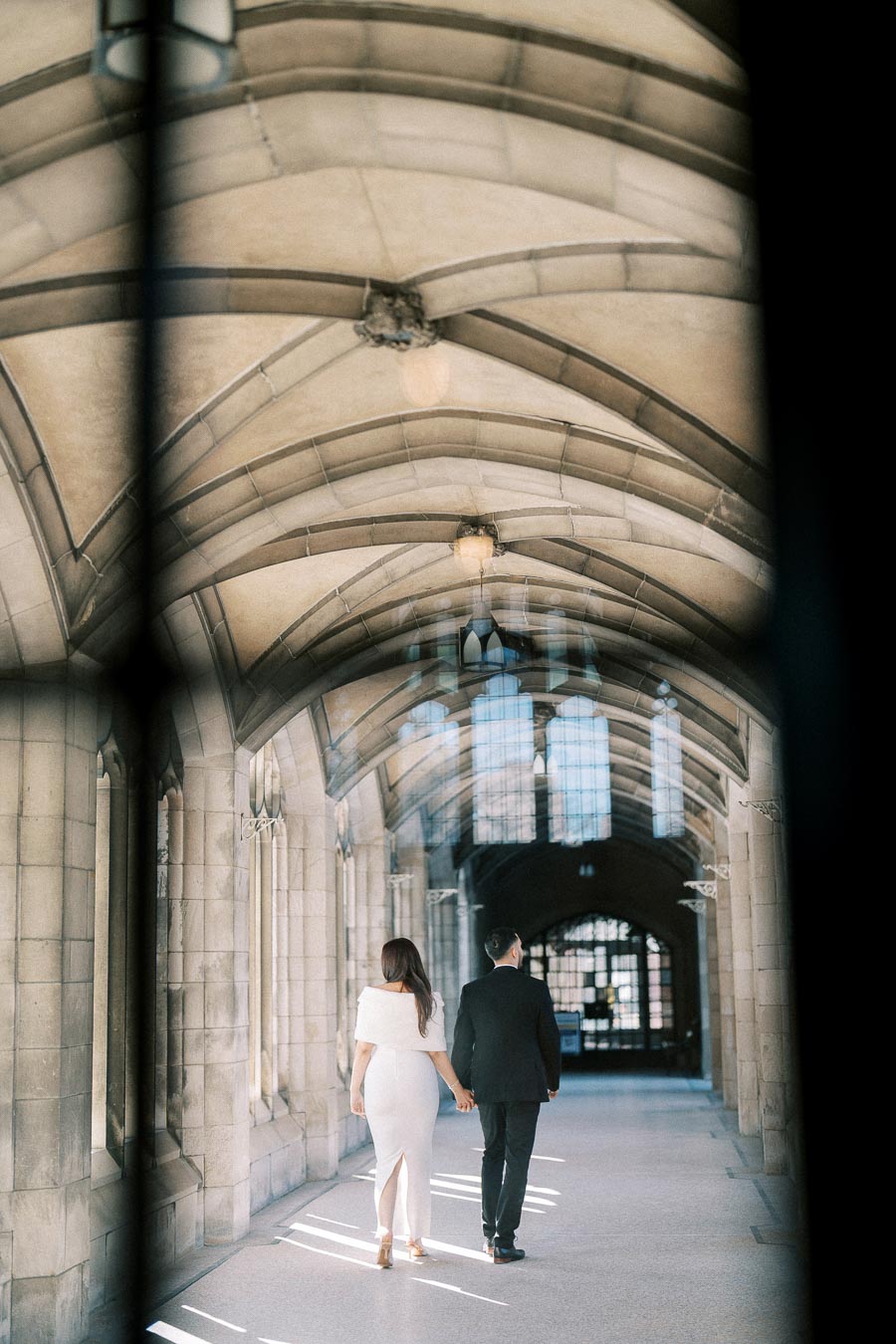 A couple holding hands walking through a sunlit, arched stone hallway, dressed in formal attire.