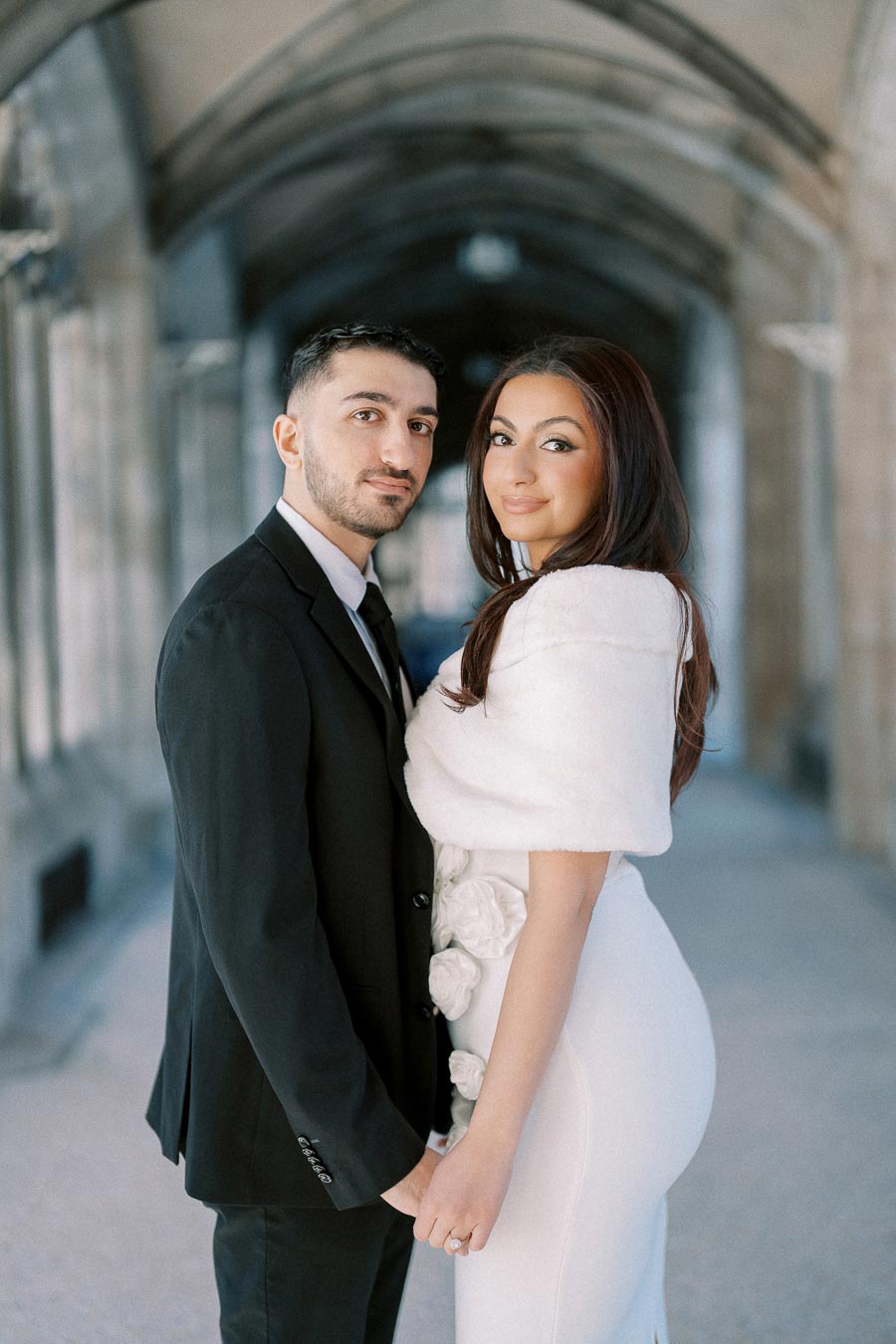 Elegant couple holding hands in a beautiful arched walkway, featuring the woman in a white dress with floral details and a