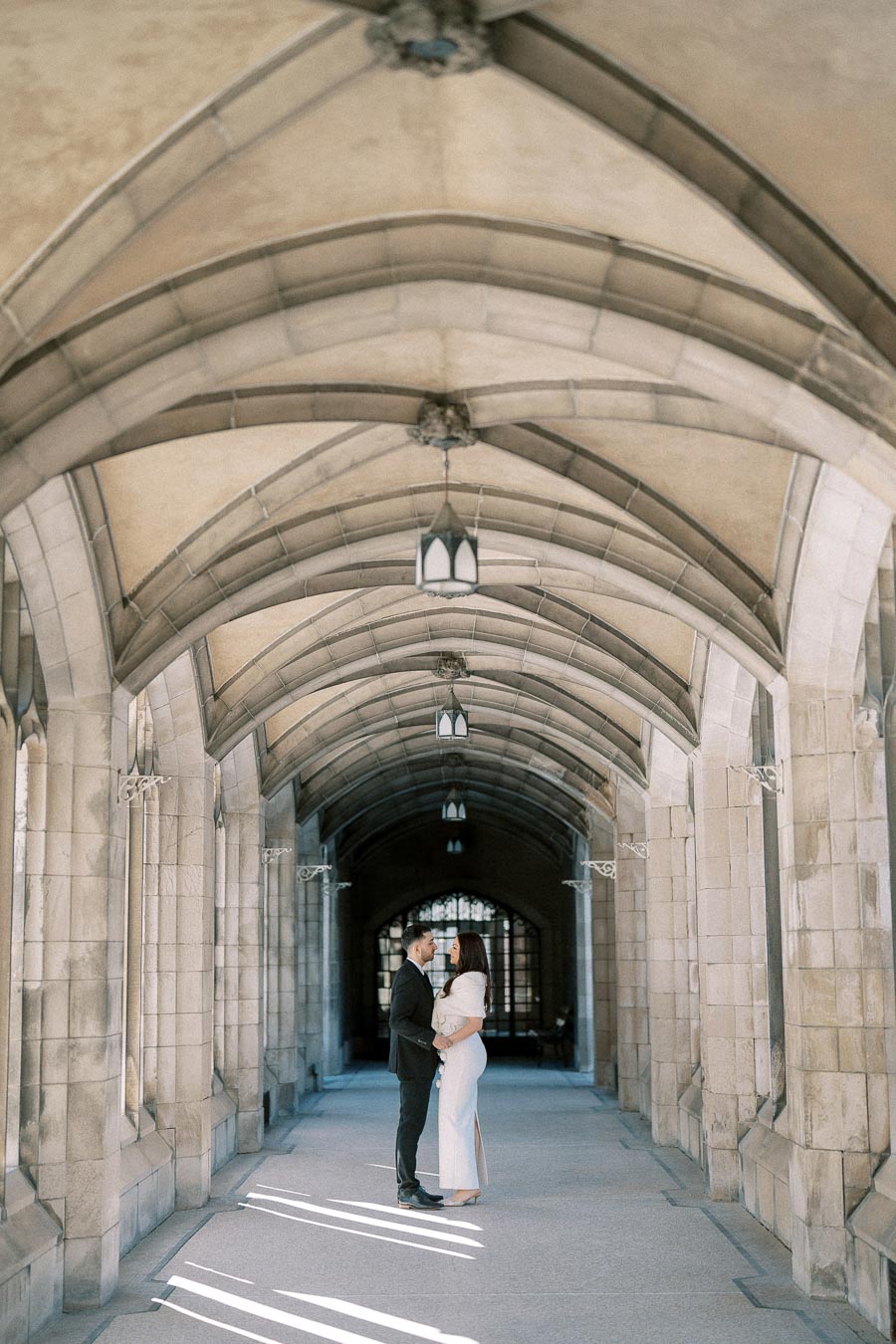 A couple embracing under a stone archway, dressed in formal attire, in a historic architectural setting.