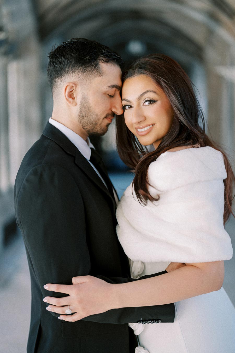A smiling couple embraces in a romantic setting, with the man wearing a black suit and the woman in a white dress and wrap,