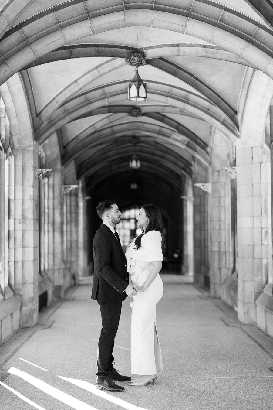 A couple smiling at each other in a beautifully arched stone corridor, dressed in formal attire, enhancing the romantic