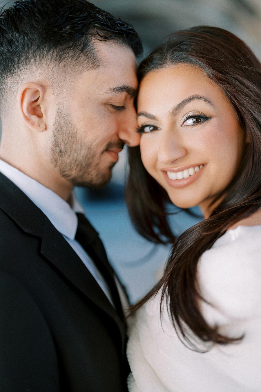 Smiling couple in elegant attire, close together in an affectionate pose, with a blurred background creating a romantic