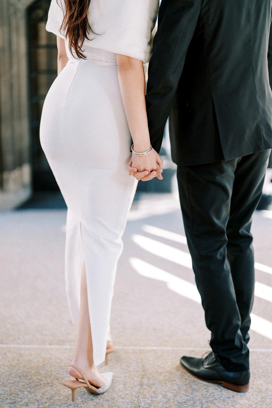 Elegant couple holding hands, woman in white dress and man in black suit, walking together, showcasing romantic attire.