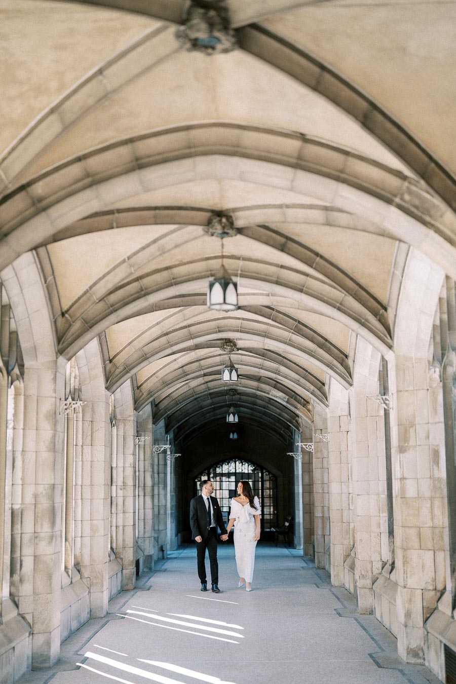 Couple holding hands in elegant stone corridor with arched ceiling, soft sunlight creating patterns on the ground.