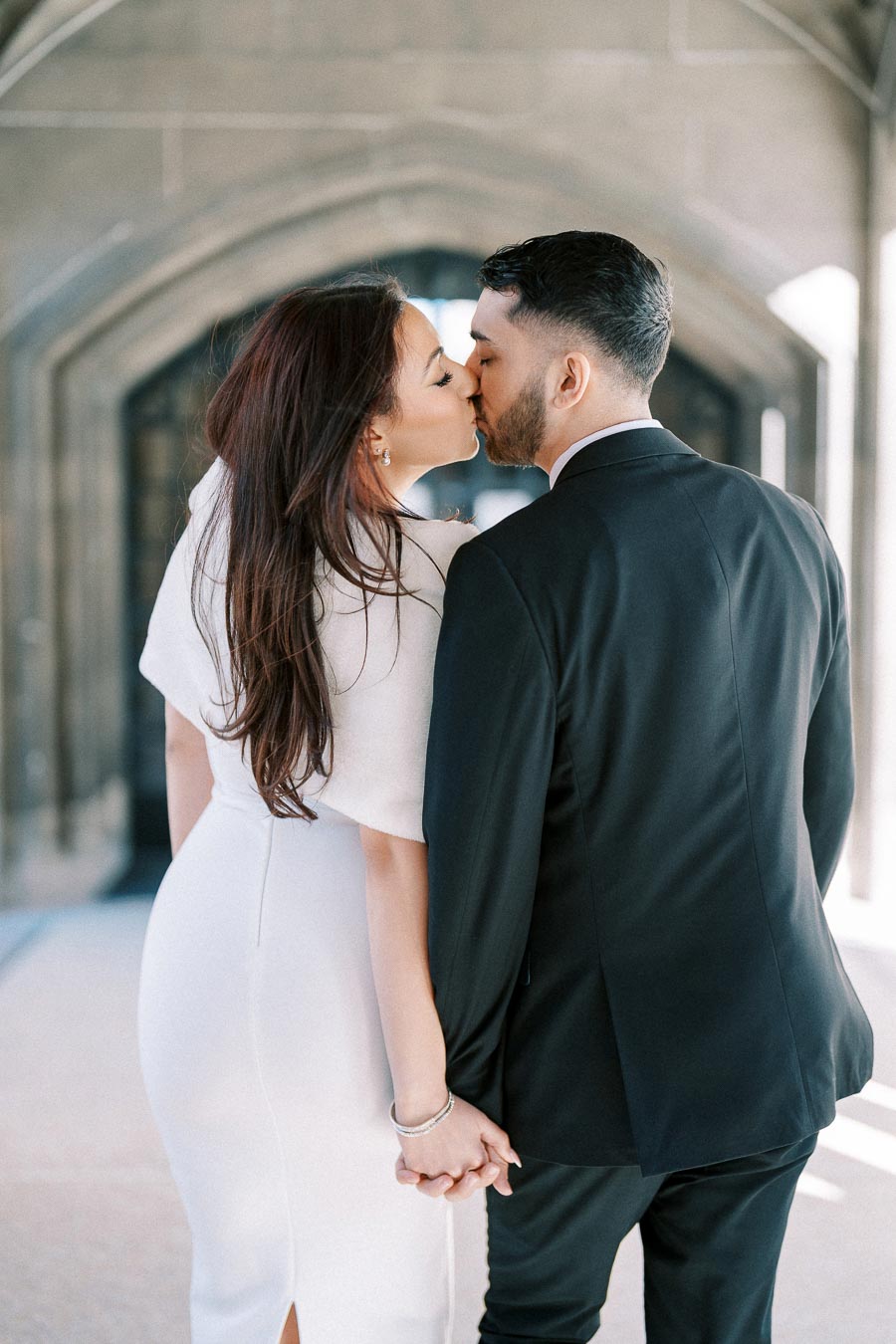 A couple dressed elegantly in formal attire shares a romantic kiss while holding hands, set against a beautiful