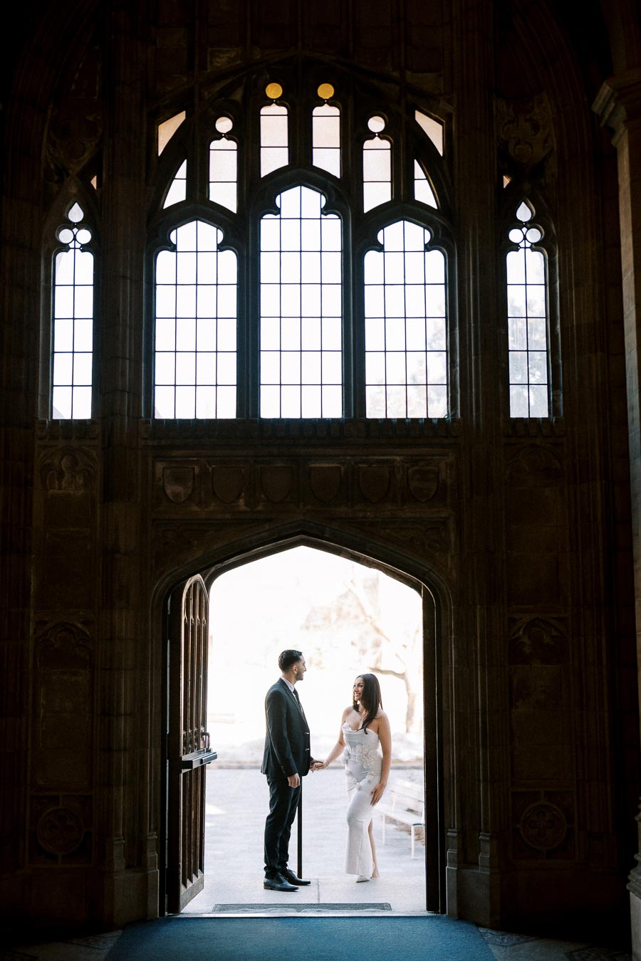 A couple holding hands in the entrance of a historic building with large arched windows, creating a romantic and elegant