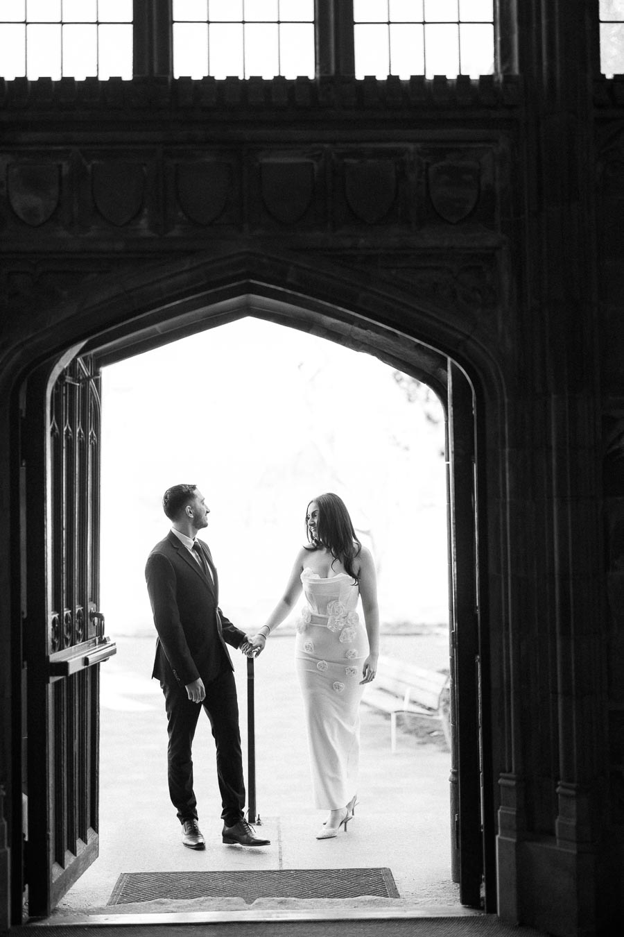 A couple holding hands under a gothic archway, wearing elegant formal attire, captured in a black and white setting.