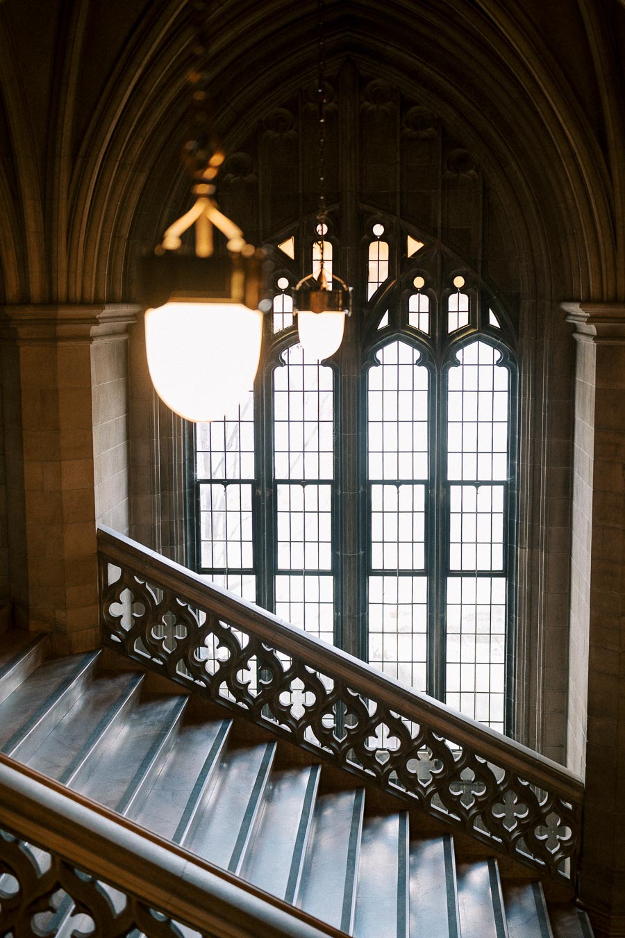 Historic stone staircase with ornate railing and glowing chandeliers inside a grand gothic building, featuring large arched
