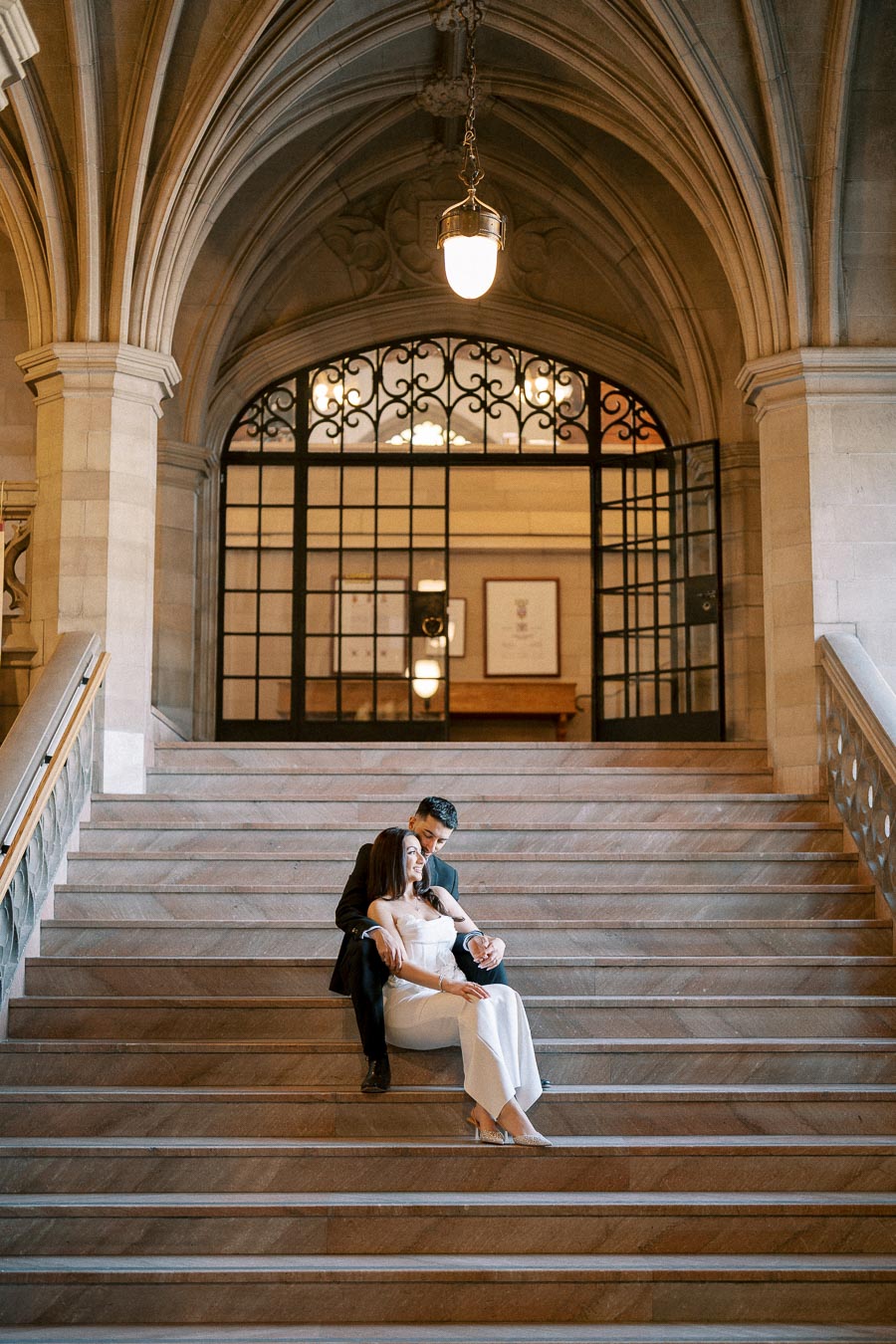 A couple sitting romantically on grand stone steps inside an elegant, historic building with arched ceilings and ornate