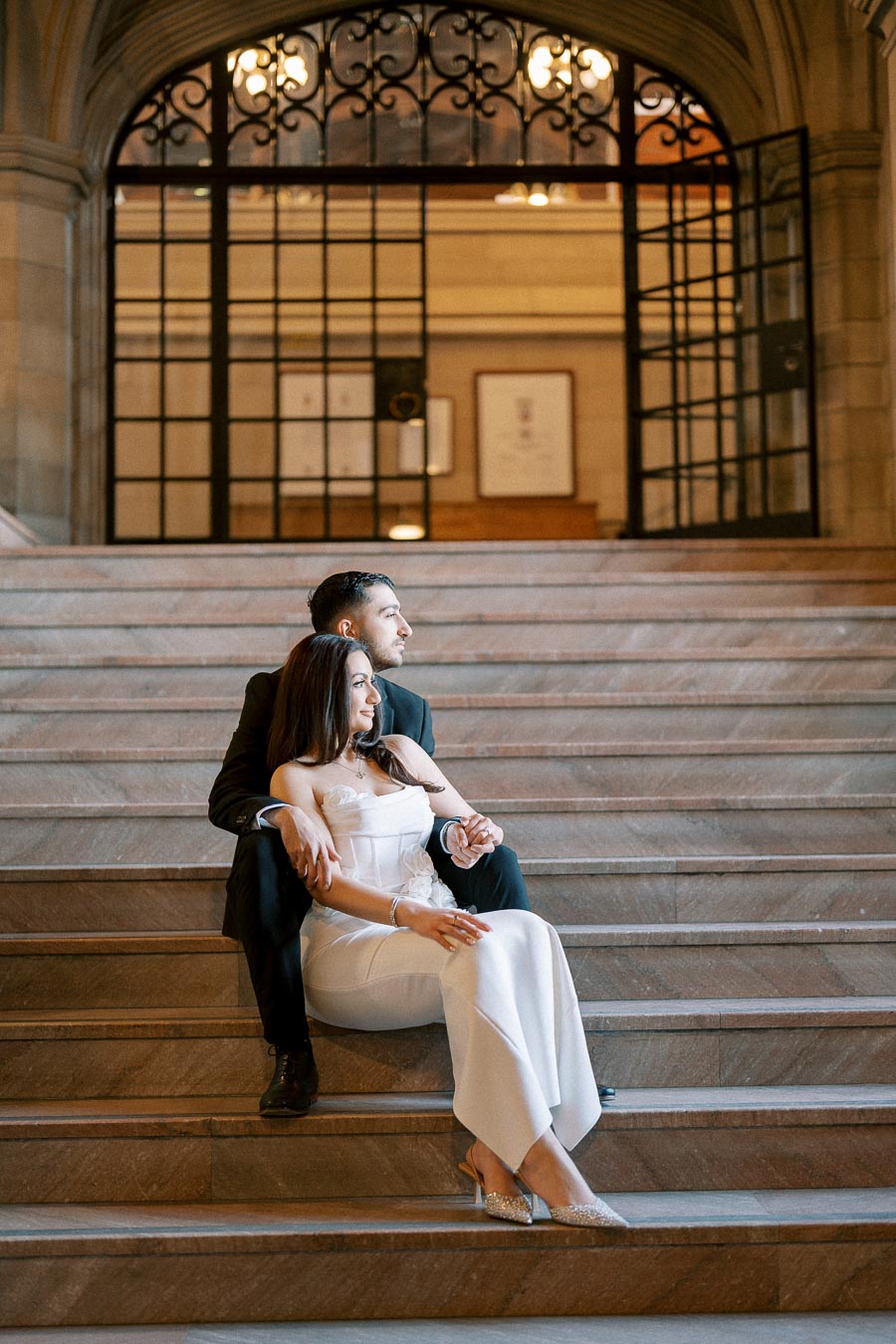 A couple in elegant attire sits on grand stone steps inside a historic building with ornate iron doors and warm lighting in