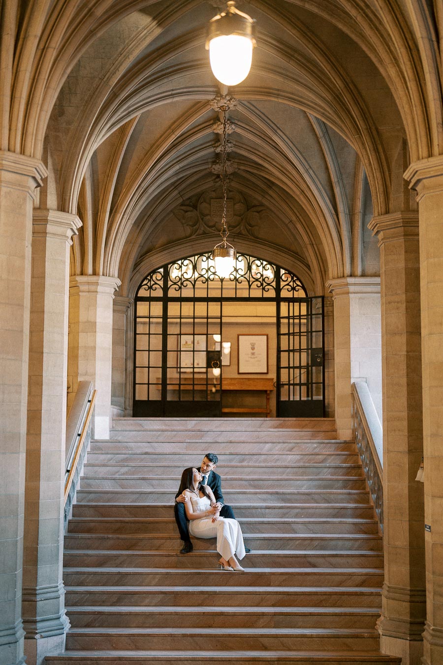 Romantic couple sitting on grand stone staircase in elegant historic building with arched ceiling and intricate ironwork in