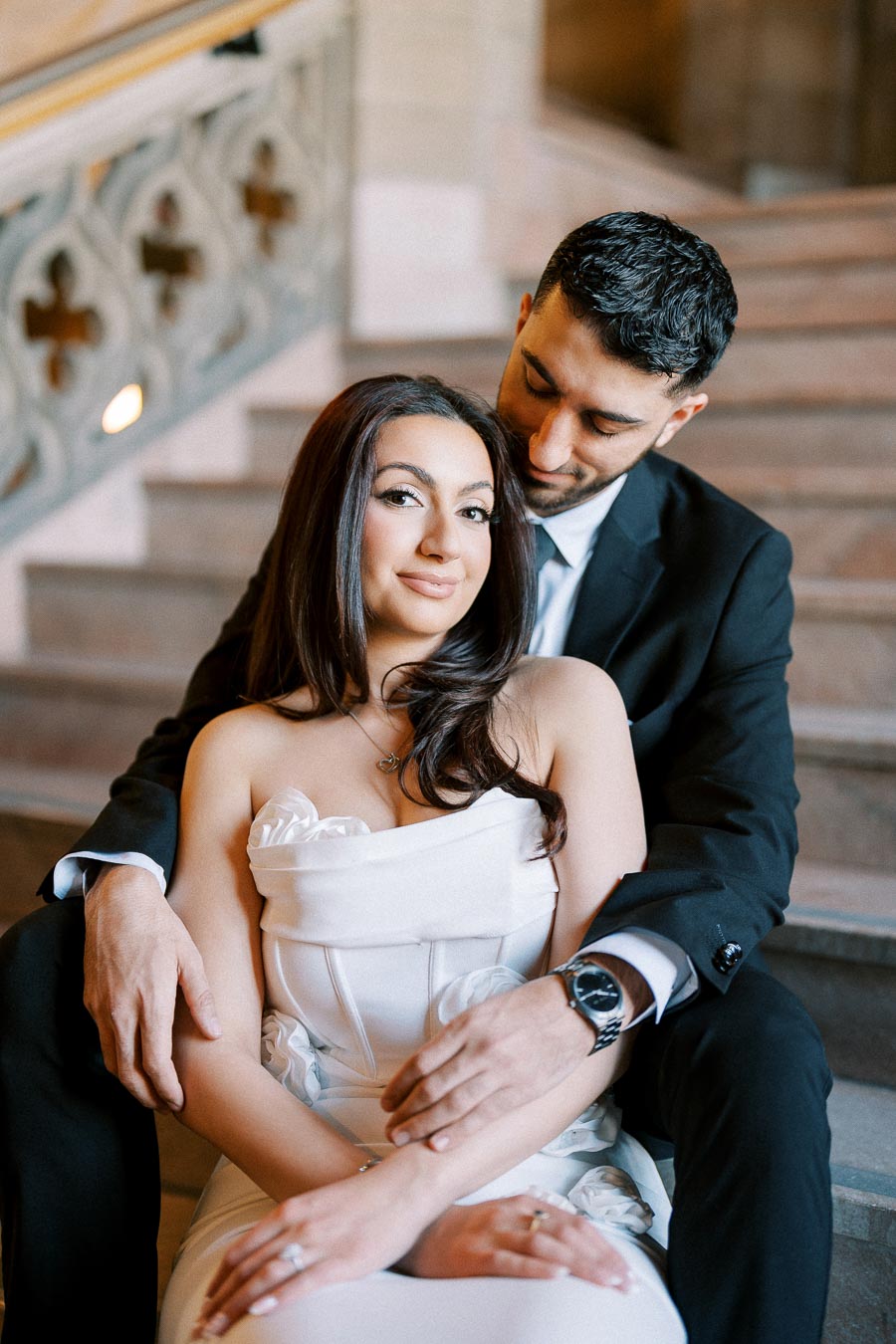 A couple sitting on stone stairs, with the woman in a white strapless dress and the man embracing her from behind in a dark