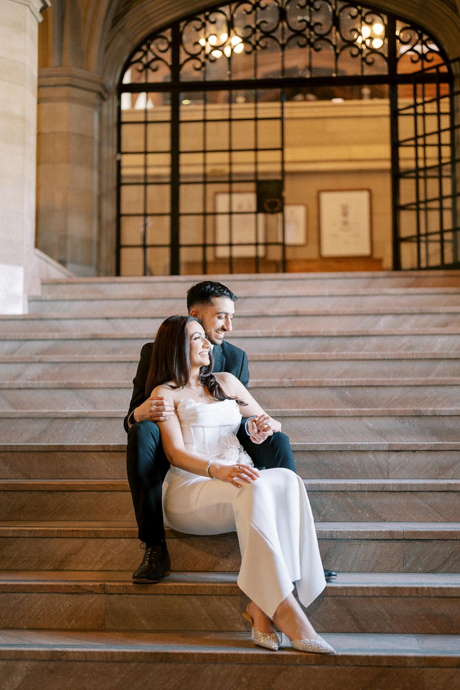 A couple sitting on elegant marble steps, smiling warmly, in a historic building with ornate ironwork and soft lighting,