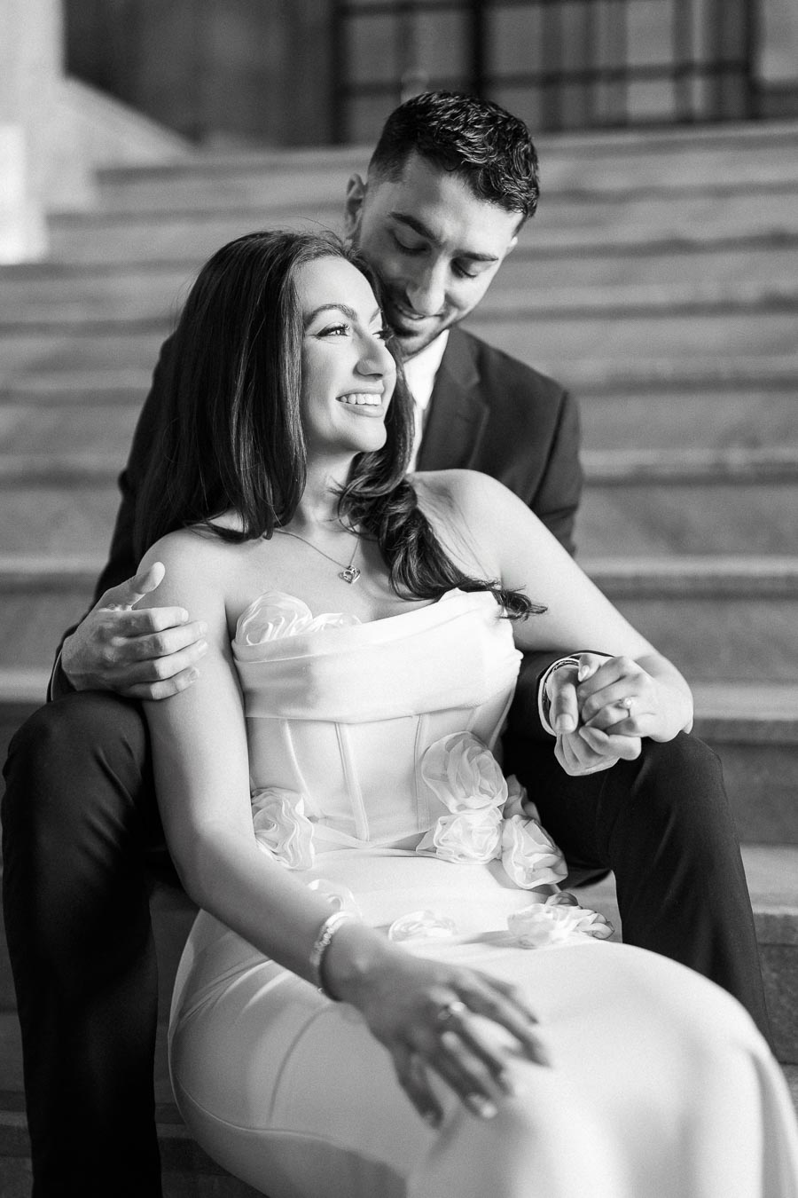 A black and white photo of a couple sitting on steps, with the woman in a white dress smiling and the man in a suit sitting