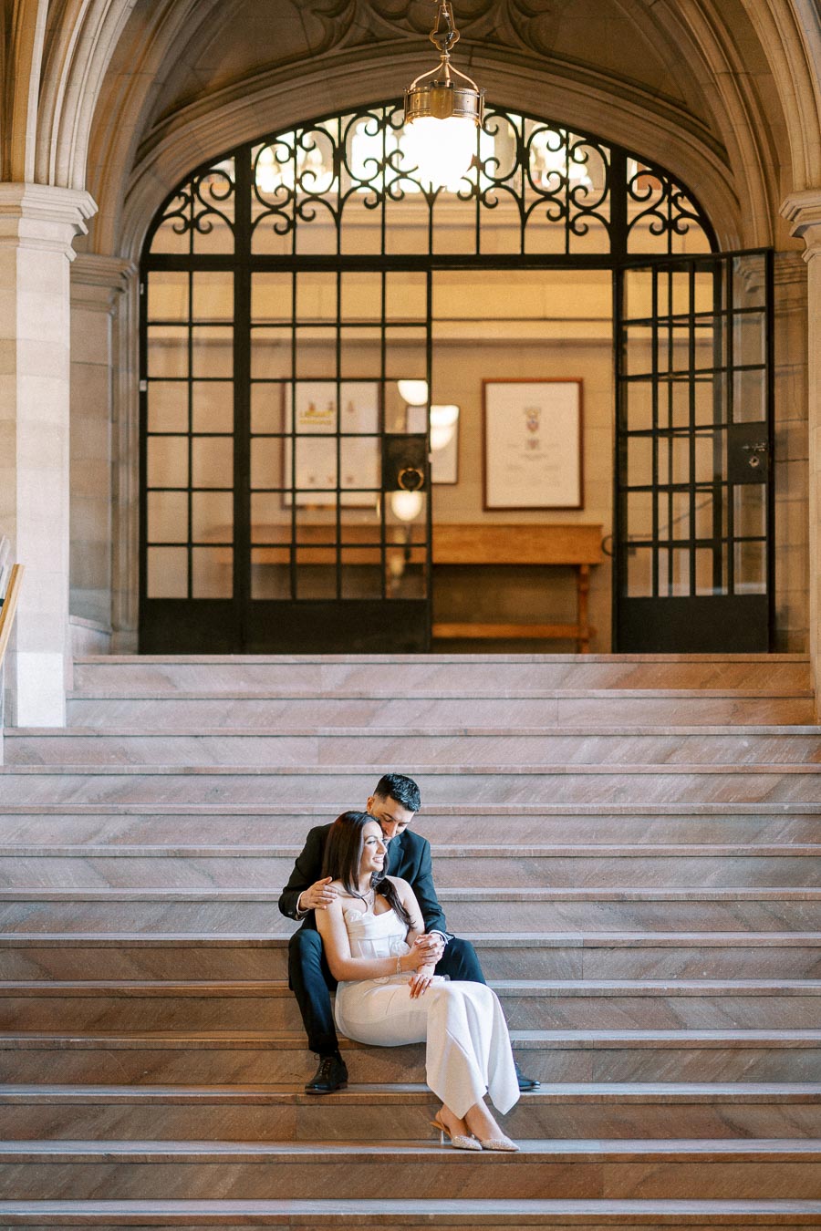 A couple in elegant attire sits closely together on grand stone steps inside a historic building, showcasing an intimate