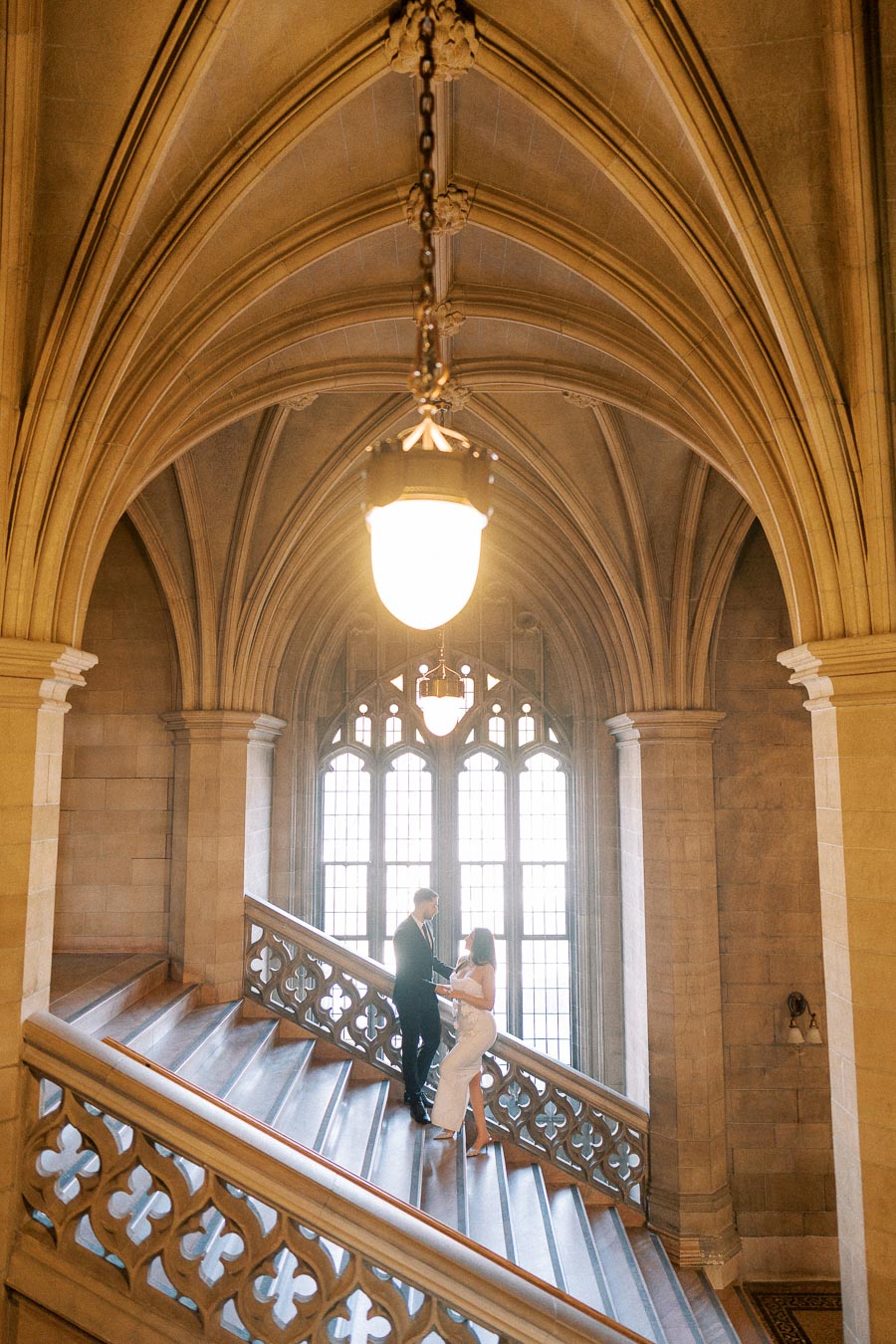 A couple embracing on a grand staircase inside a Gothic-style building with arched ceilings and ornate stonework,