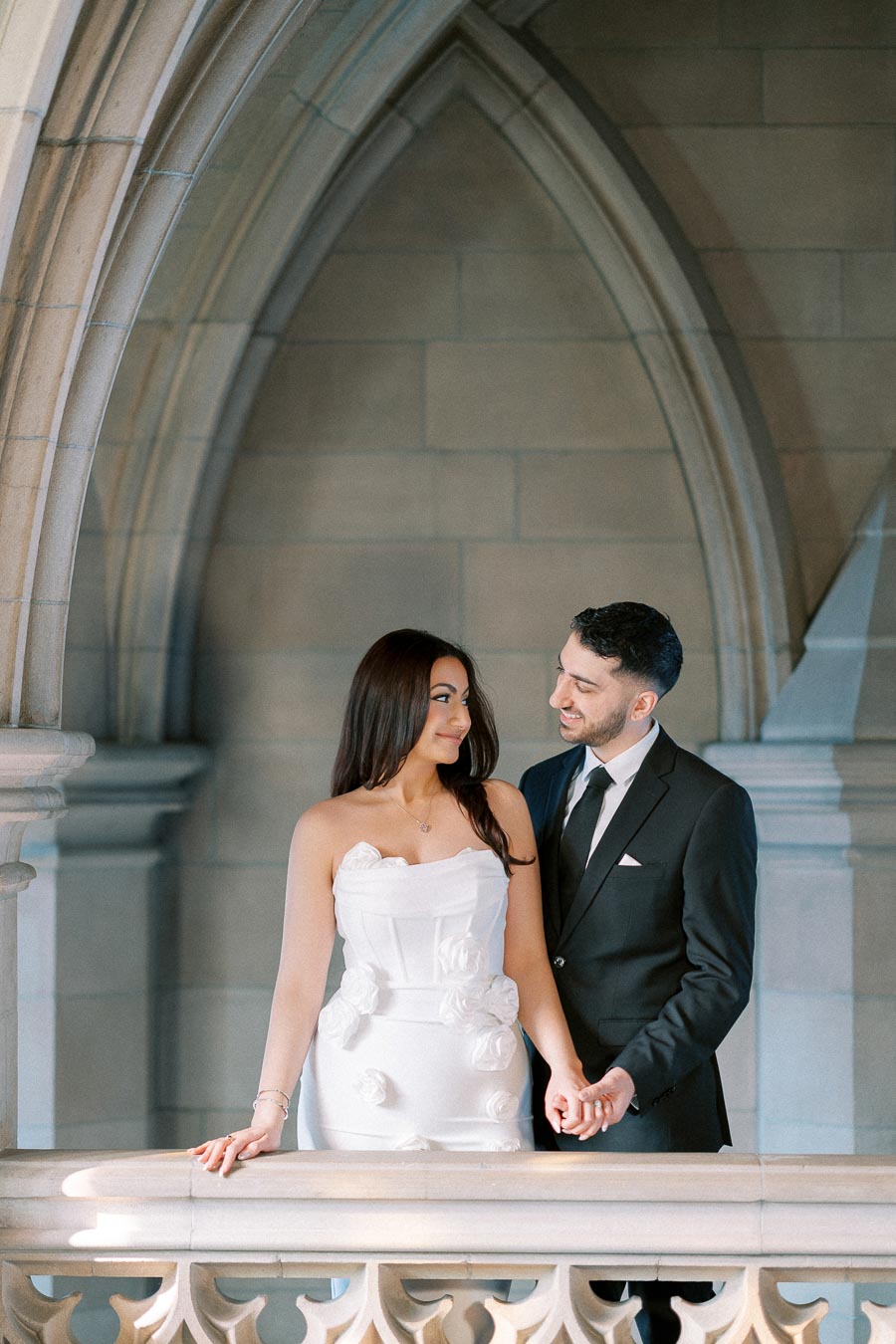 Elegant couple standing on balcony with gothic architecture backdrop, woman in white dress with floral details and man in