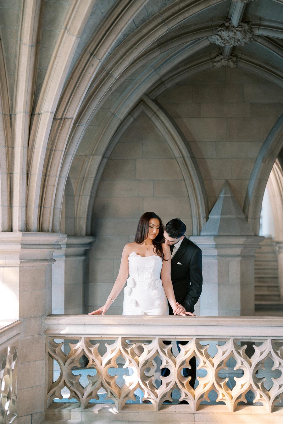 Elegant couple in formal attire posing in a historic architectural setting with gothic arches and intricate stonework.