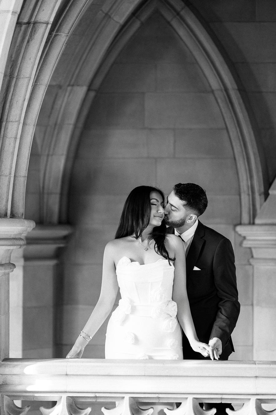 Black and white photo of a couple embracing in a romantic moment under an elegant stone archway, the woman wearing a