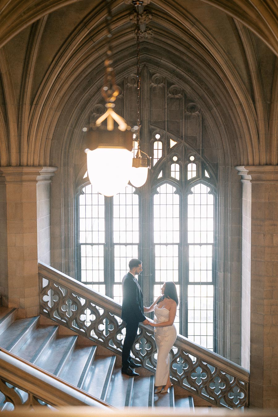 Couple elegantly dressed, standing on a grand staircase within a historic Gothic-style building, featuring arched windows