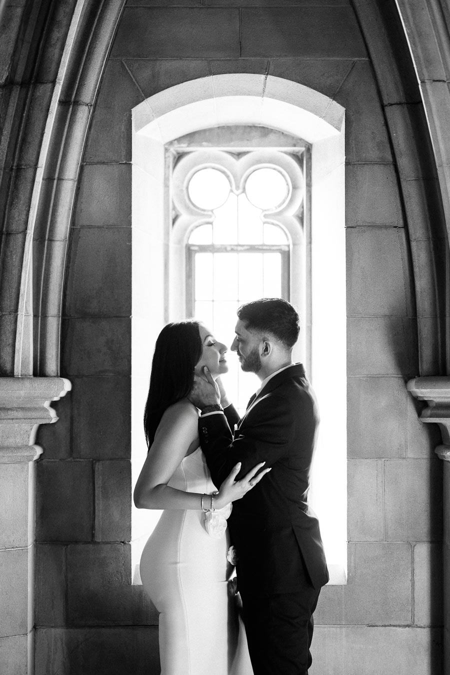 A black and white photograph of a couple in formal attire sharing a romantic moment in a stone archway, with light streaming