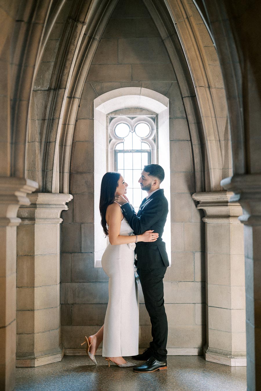 A romantic couple embracing inside a gothic-style stone archway, with soft natural light streaming through a decorative