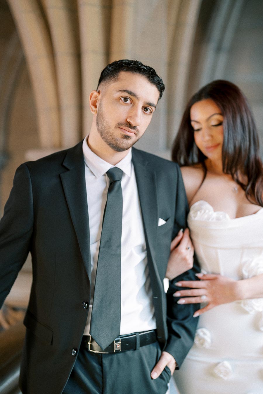 A man in a black suit and tie stands next to a woman in a white dress with floral details, set against an elegant