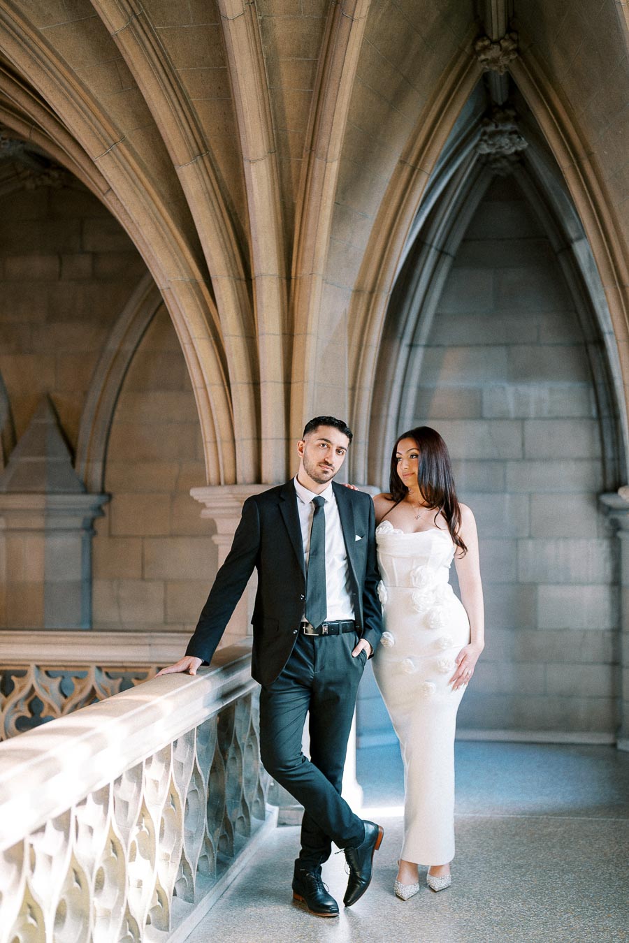 A couple elegantly dressed, posing in a historic architectural setting with stone archways and intricate railing designs.