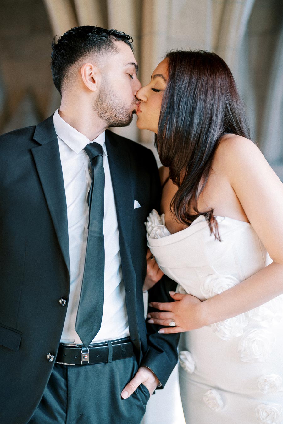 A couple dressed elegantly in a black suit and white dress shares a tender kiss in a romantic setting.
